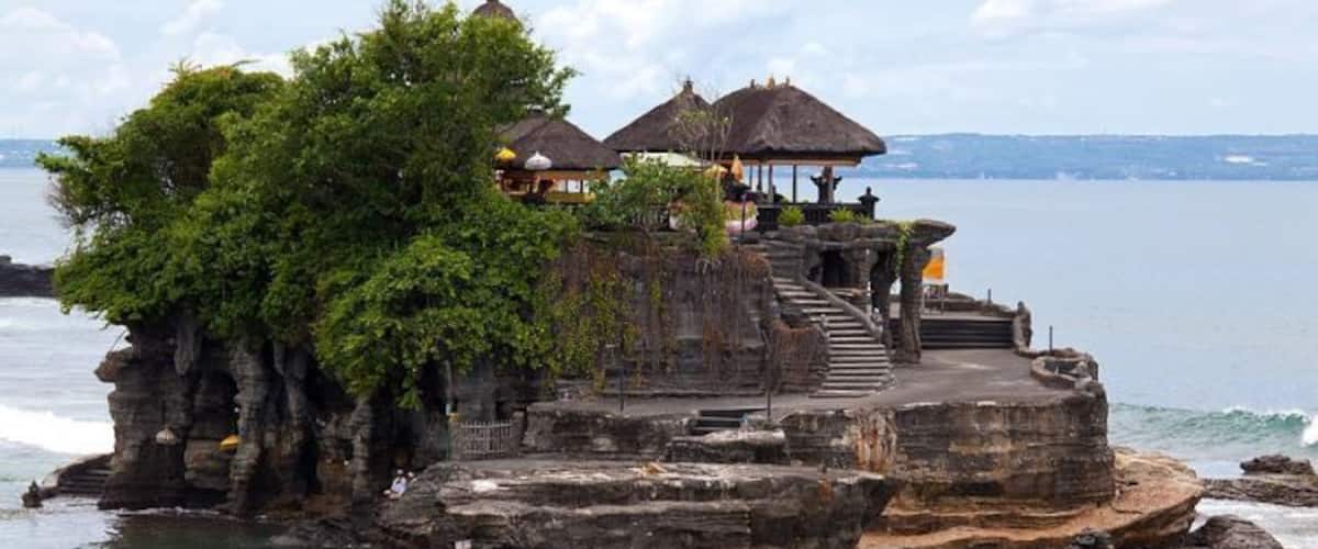 A stone structure with a thatched roof and stairs leading up to a platform, surrounded by lush greenery and overlooking a body of water.