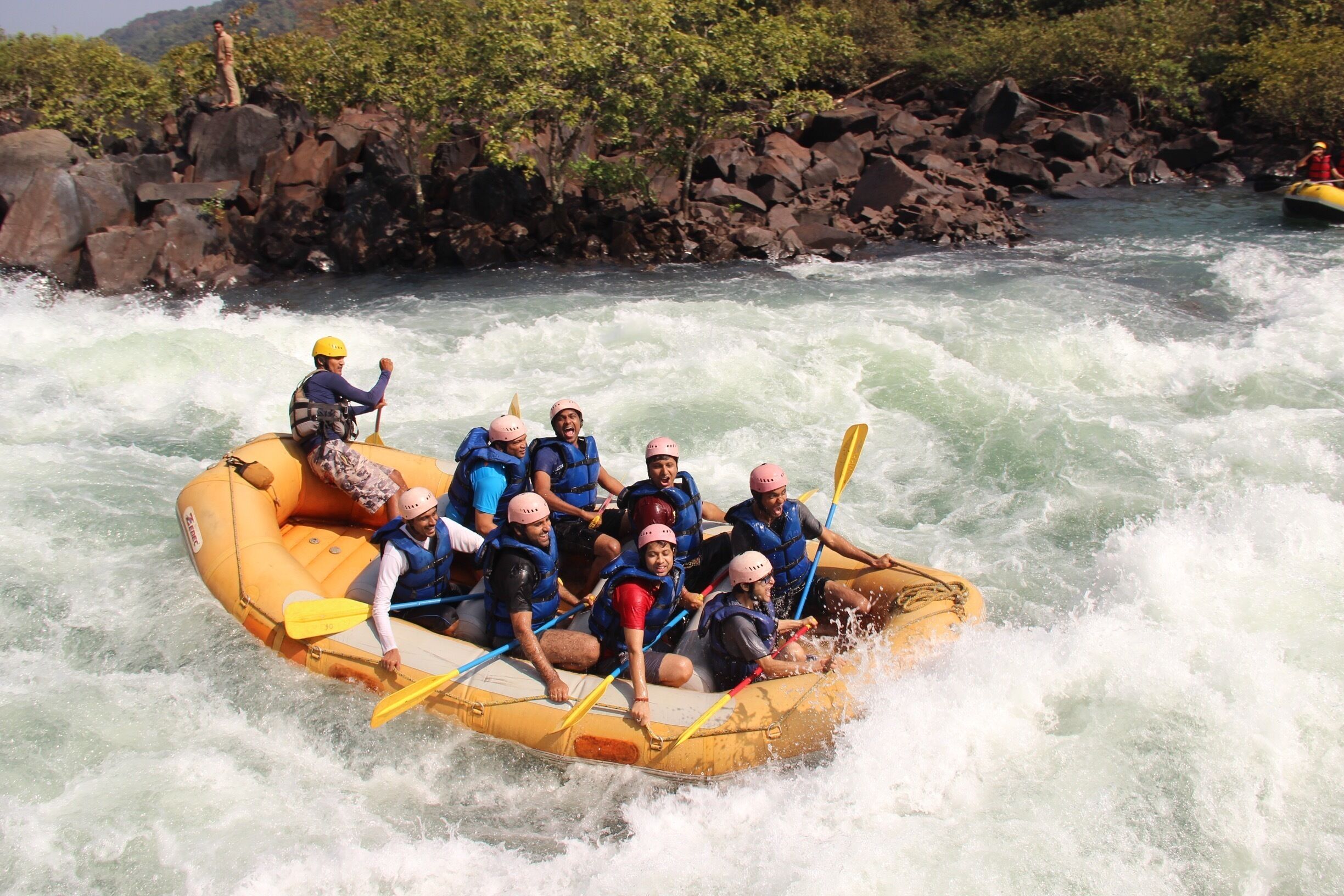 A group of people in a yellow raft navigating through a river with rapids, surrounded by rocky terrain and greenery.
