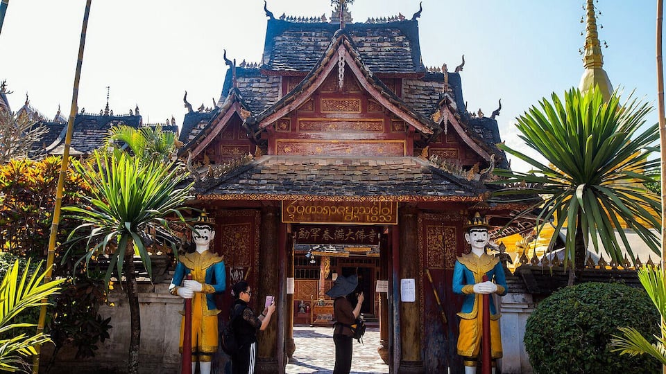 A traditional Thai temple with ornate architecture, including a multi-tiered roof and golden statues at the entrance.