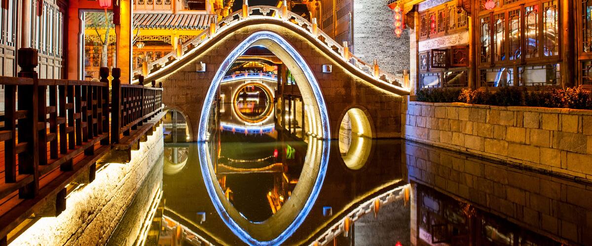 Ein traditioneller chinesischer Wasserweg mit einer kreisrunden Steinbrücke, die die umliegenden Gebäude und Laternen spiegelt.