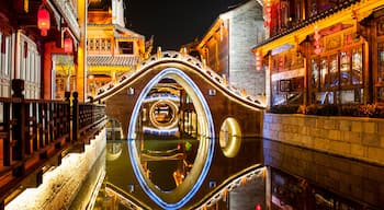 Ein traditioneller chinesischer Wasserweg mit einer kreisrunden Steinbrücke, die die umliegenden Gebäude und Laternen spiegelt.