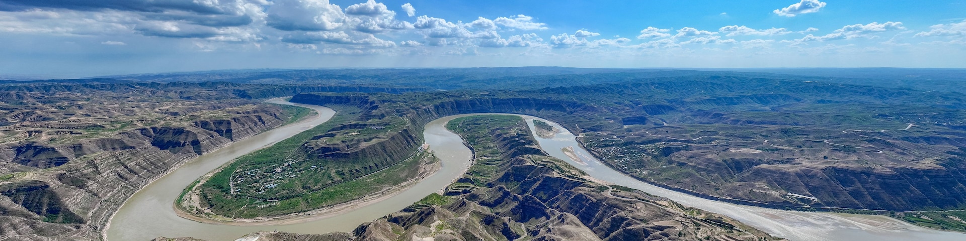 Um rio serpenteia por um cânion acidentado com vegetação escassa e edifícios ao longe.