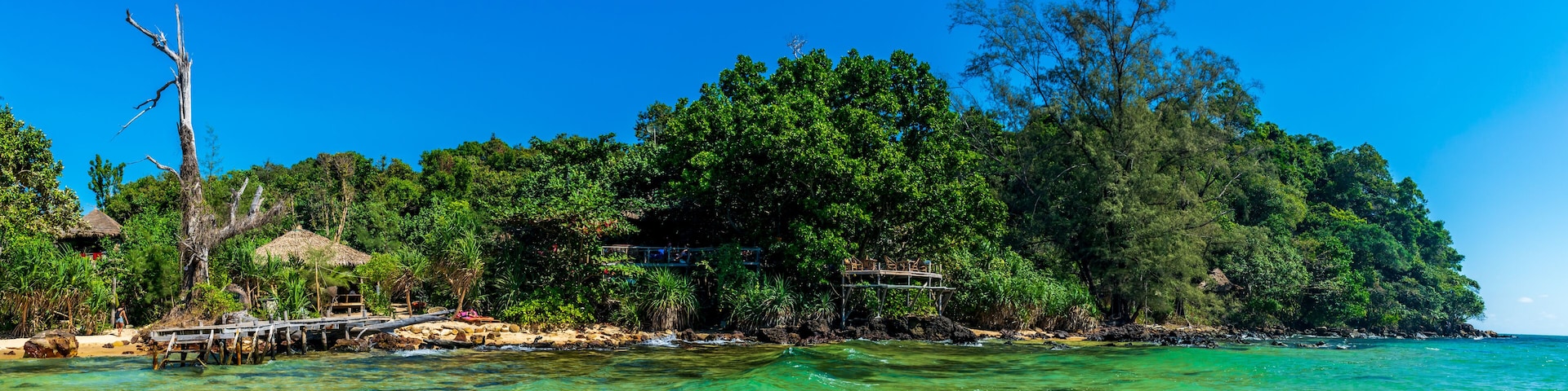 A tropical island with clear turquoise waters, a wooden pier, lush greenery, and a thatched-roof structure.