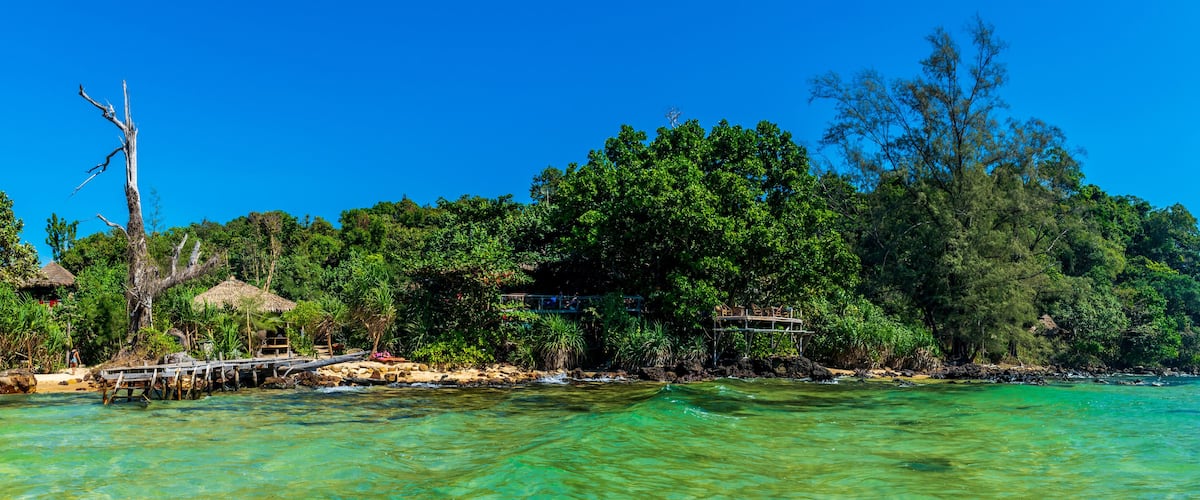 A tropical island with clear turquoise waters, a wooden pier, lush greenery, and a thatched-roof structure.