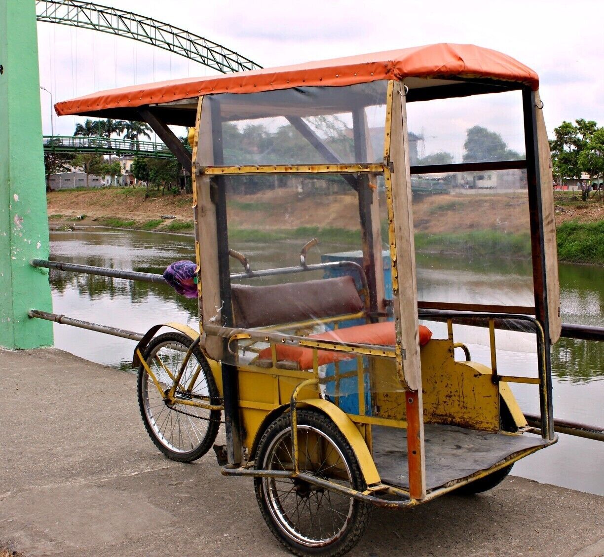A yellow and red rickshaw parked by a canal with a green bridge in the background.