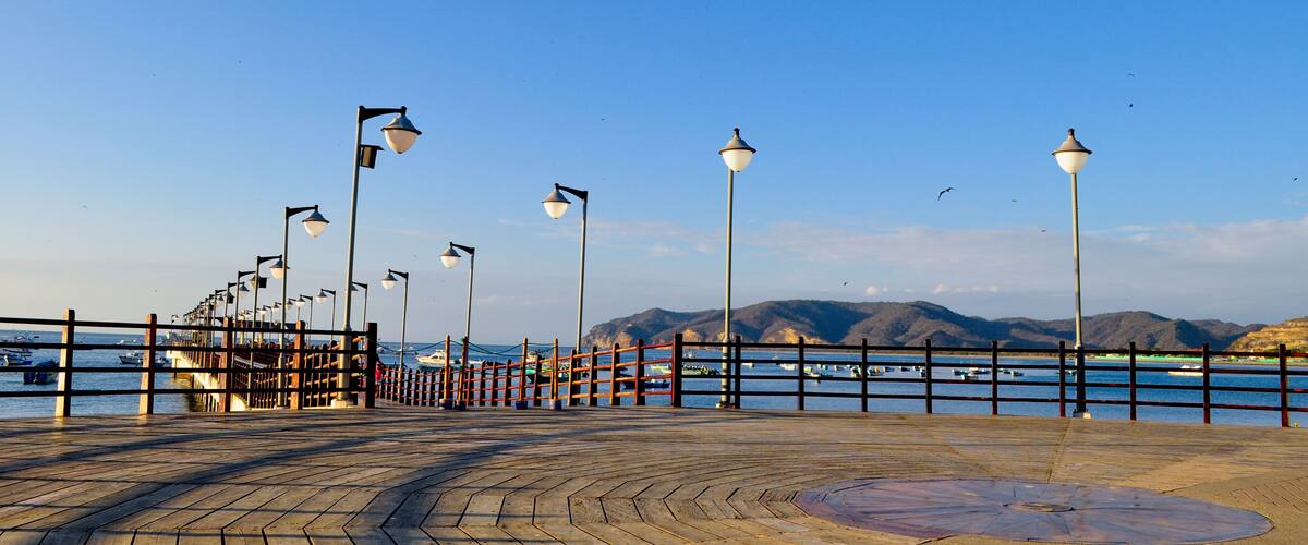 A wooden boardwalk with street lamps and a view of boats and mountains in the distance.