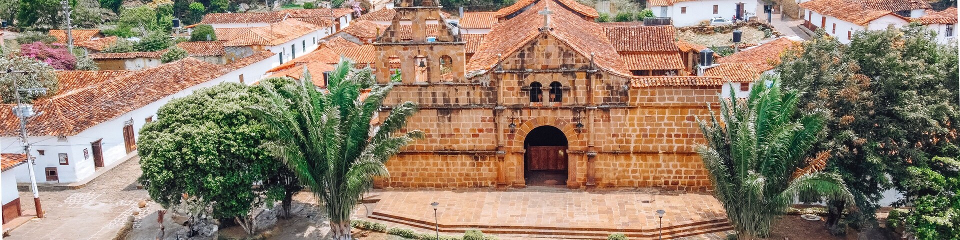 A historic church with a central archway, surrounded by white buildings with red-tiled roofs, set in a hilly area with lush greenery.