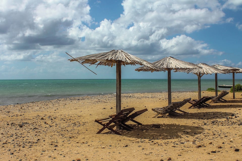 Una playa con sombrillas de paja y sillones de madera.