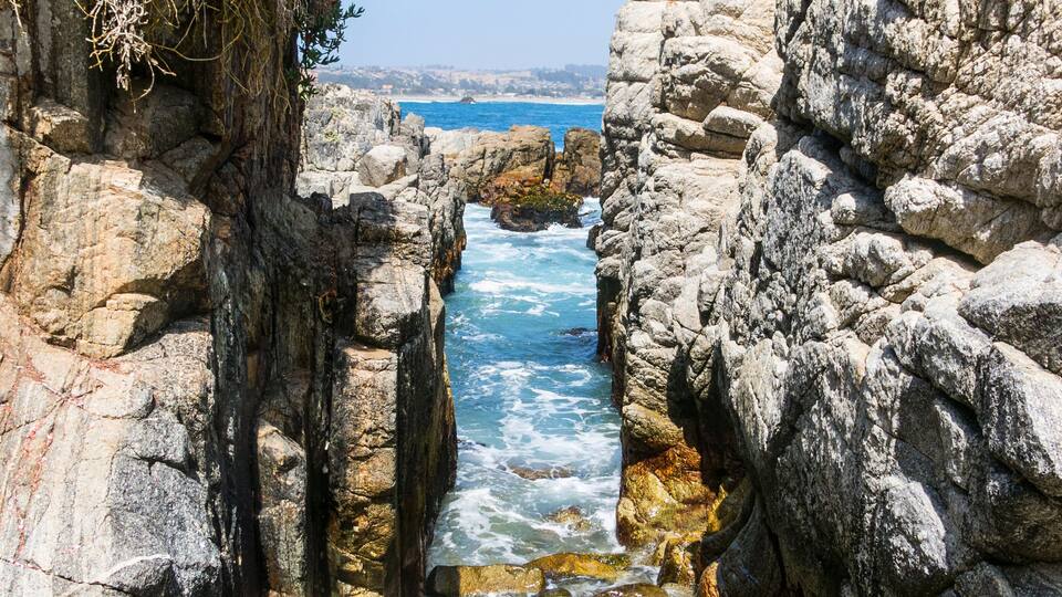 A rocky coastal path with waves crashing through a narrow gap between large boulders, leading to the sea.