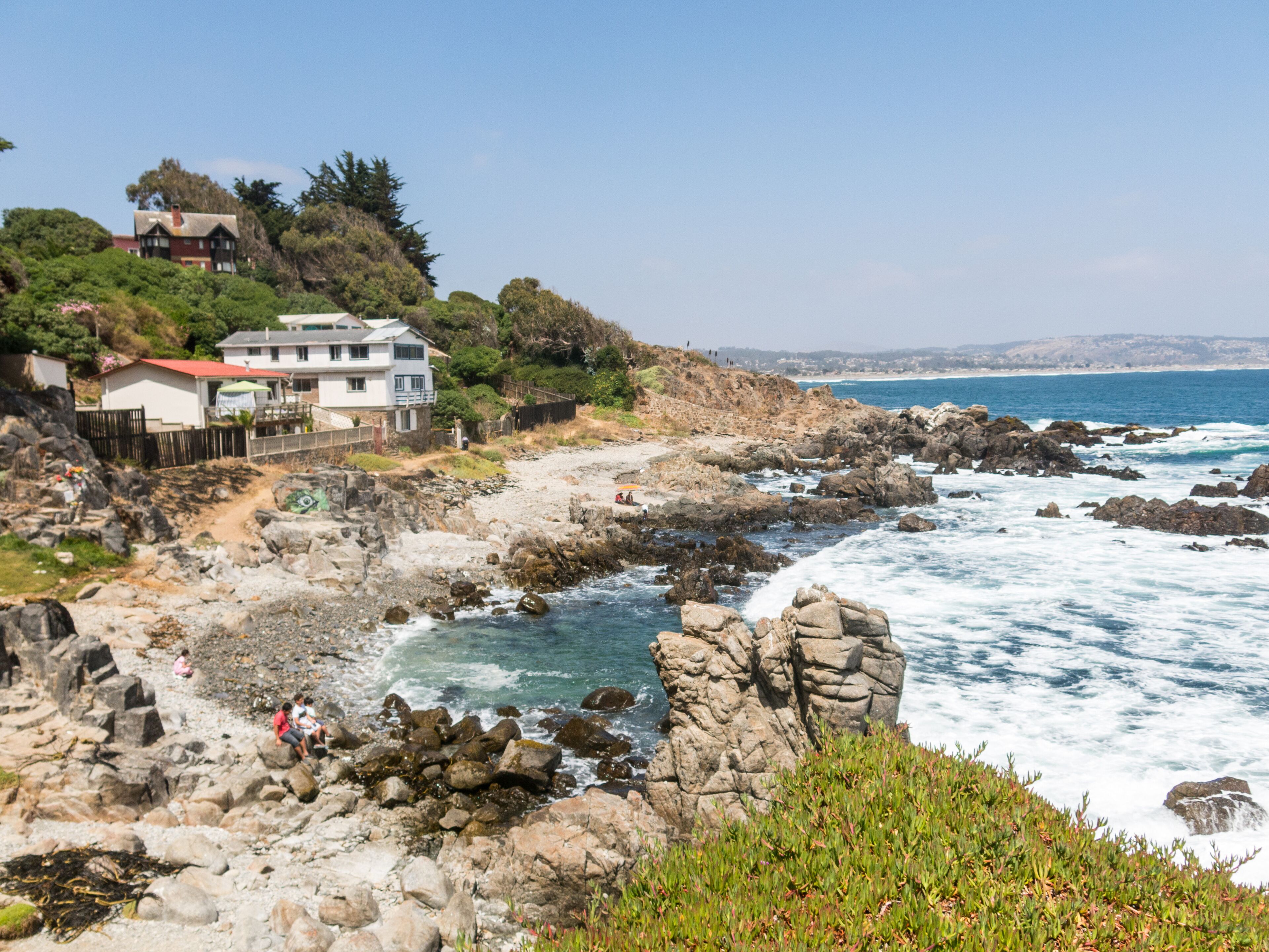A rocky coastline with waves crashing against the shore, a white house with a red roof, and lush greenery.