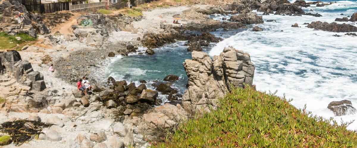 A rocky coastline with waves crashing against the shore, a white house with a red roof, and lush greenery.