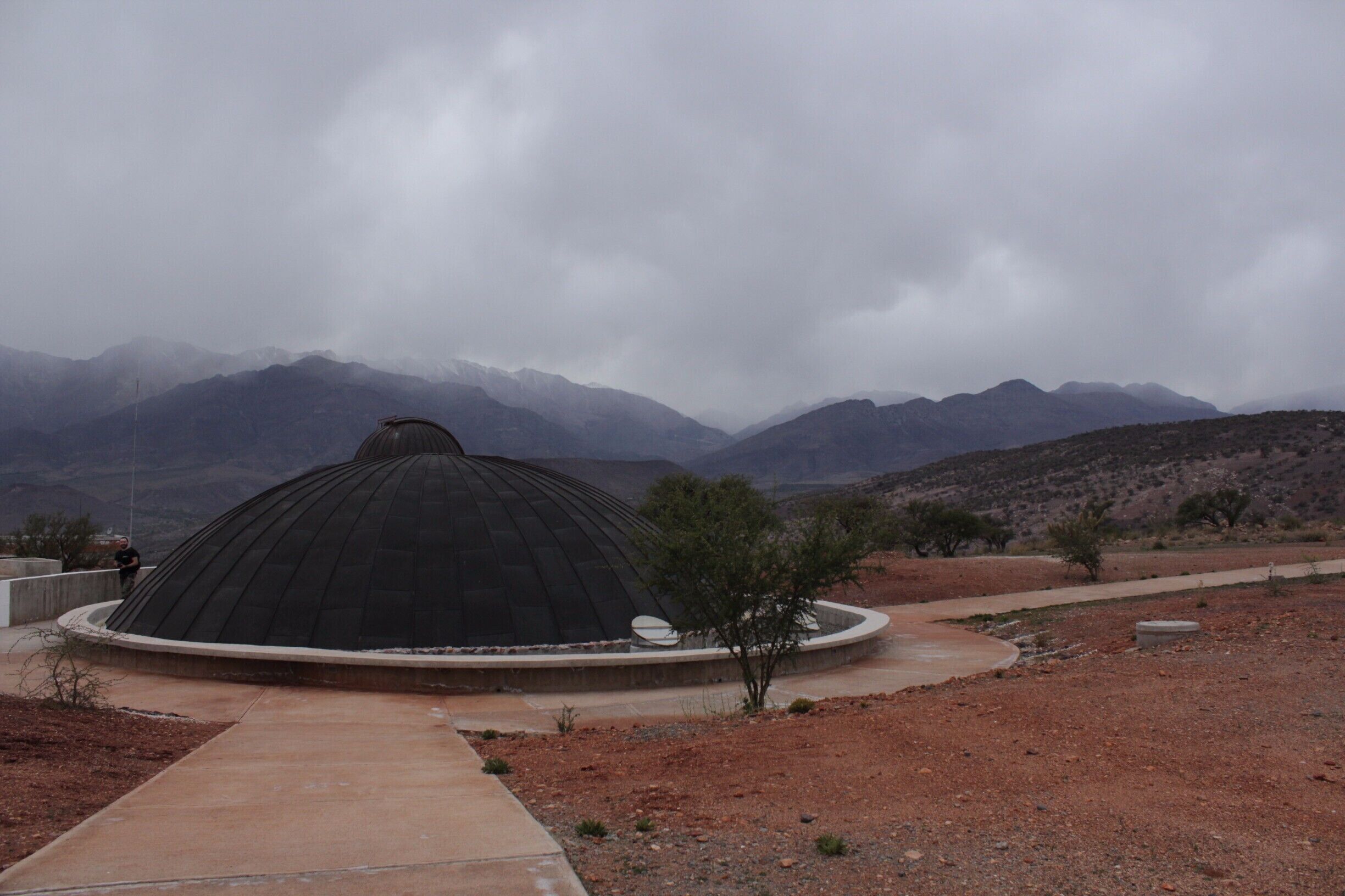 A large, dark dome structure in a desert landscape with mountains in the background.