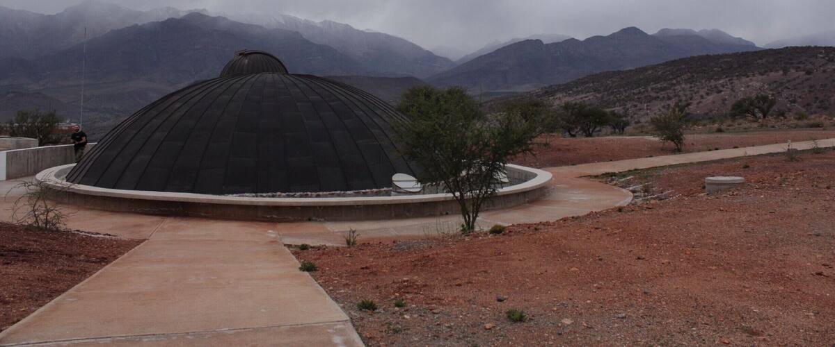 A large, dark dome structure in a desert landscape with mountains in the background.