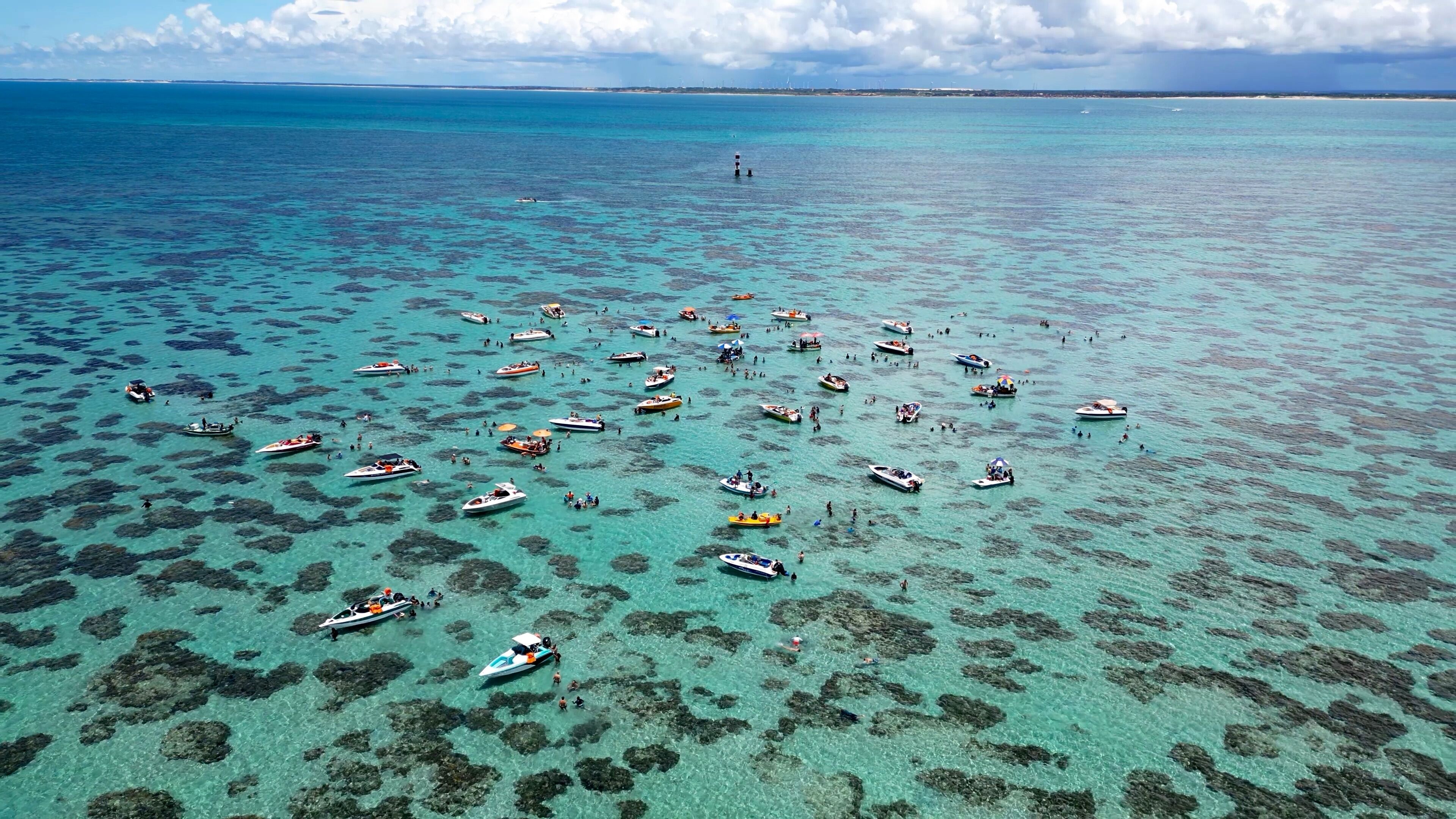 An aerial view of a turquoise sea dotted with numerous boats and people, with a distant shoreline and a lighthouse.