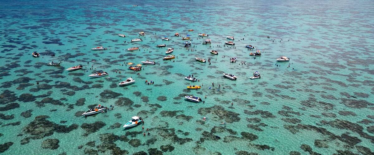 An aerial view of a turquoise sea dotted with numerous boats and people, with a distant shoreline and a lighthouse.