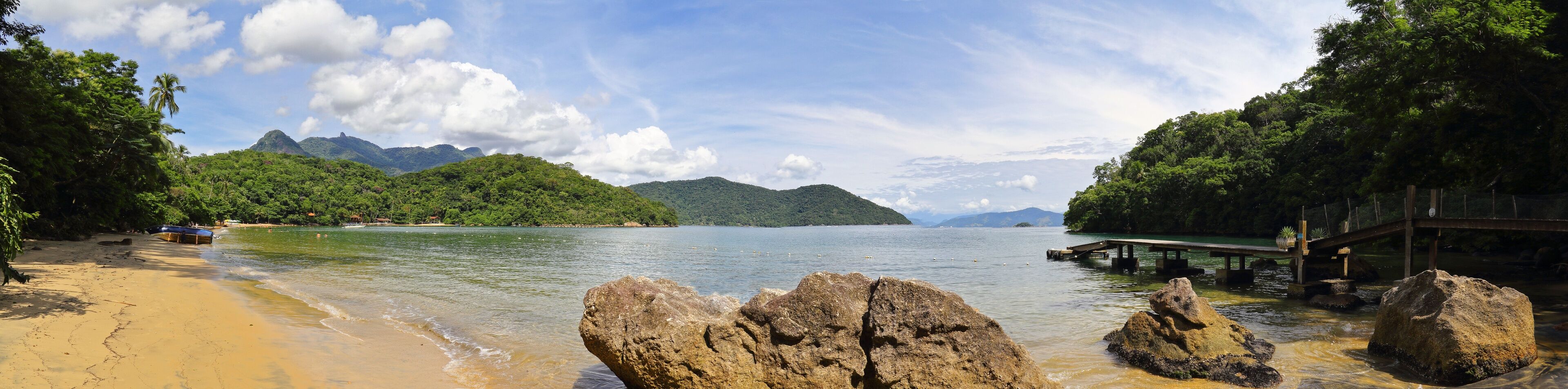 A beach with clear water, rocky outcrops, and lush green hills in the background.