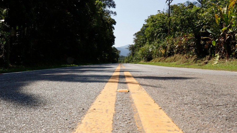 Uma estrada de duas pistas com marcação central amarela, cercada por vegetação densa e árvores.