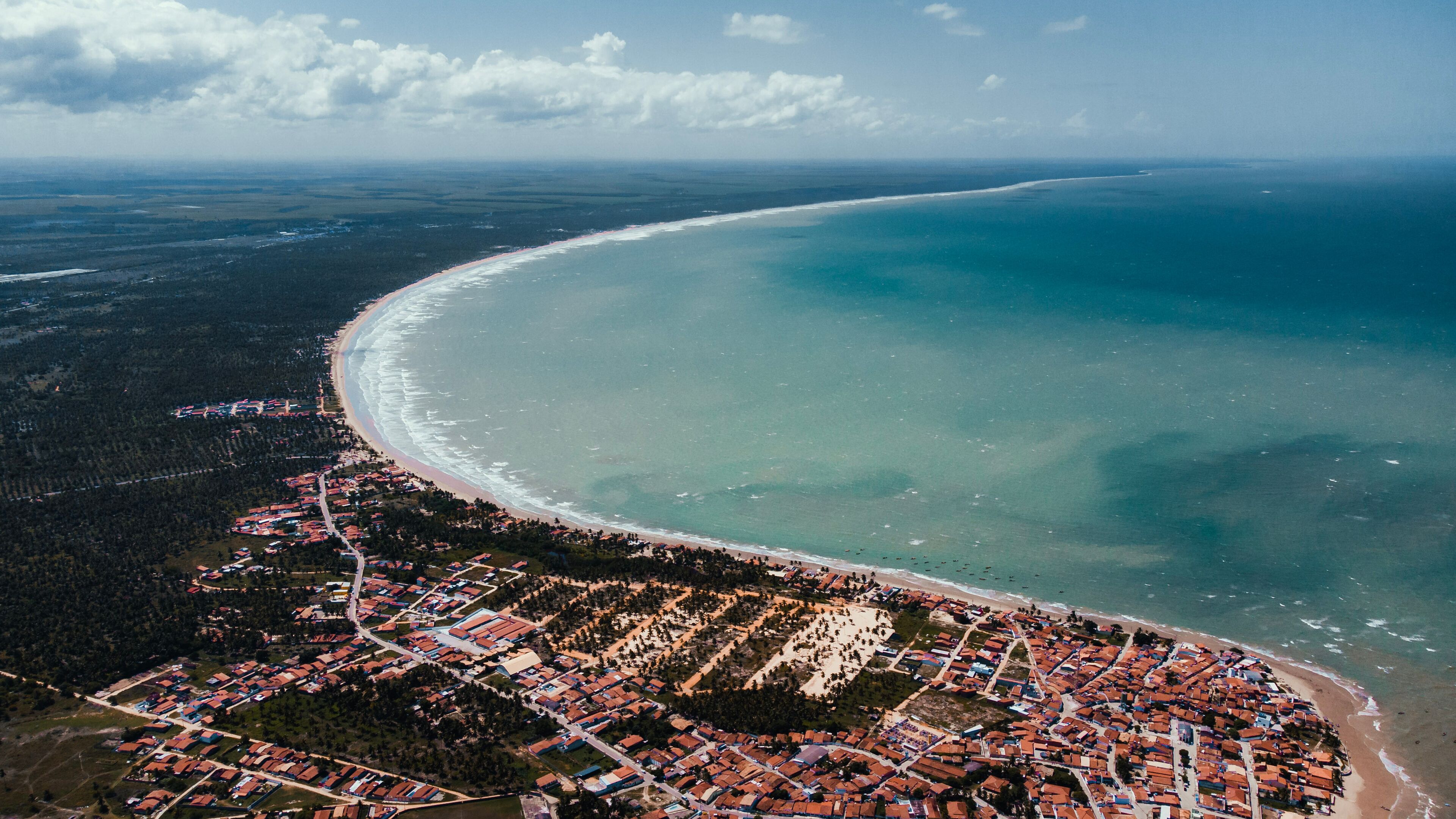 An aerial view of a coastal town with a curved beach, dense forested areas, and a clear sky.