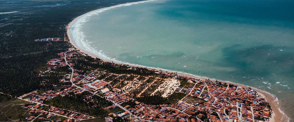An aerial view of a coastal town with a curved beach, dense forested areas, and a clear sky.