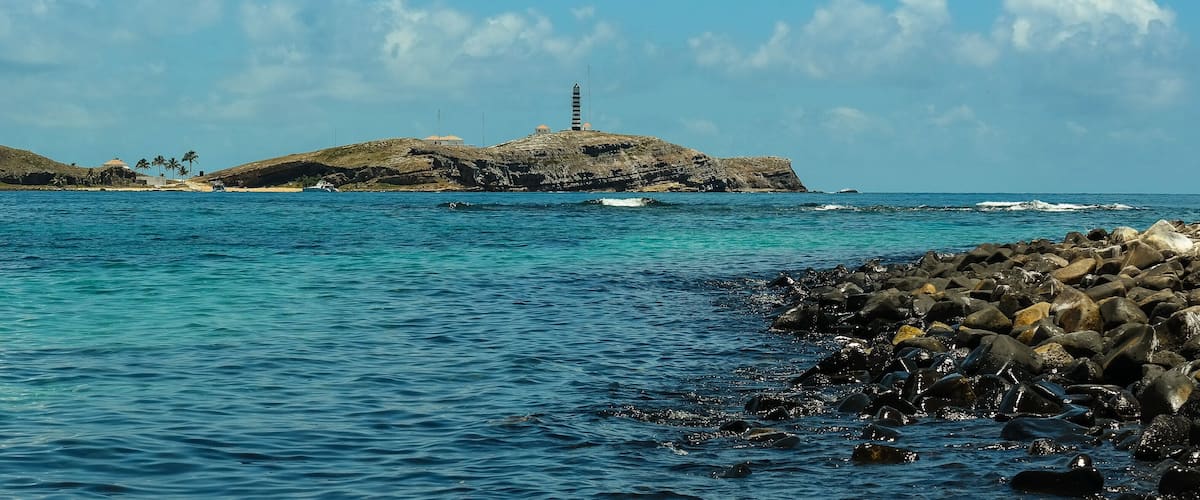 A coastal landscape with a lighthouse on a rocky island, calm sea, and clear blue sky.