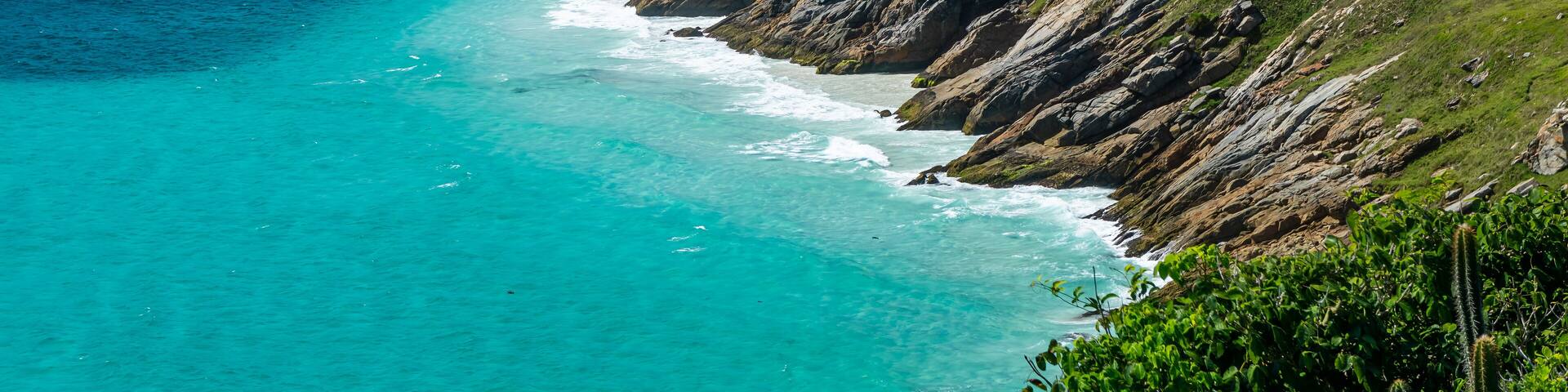 View of Guriri cove turquoise waters surrounded by Pontal do Atalaia mountains fully covered by lush dense green vegetation under summer morning blue sky in Arraial do Cabo, Rio de Janeiro - Brazil.