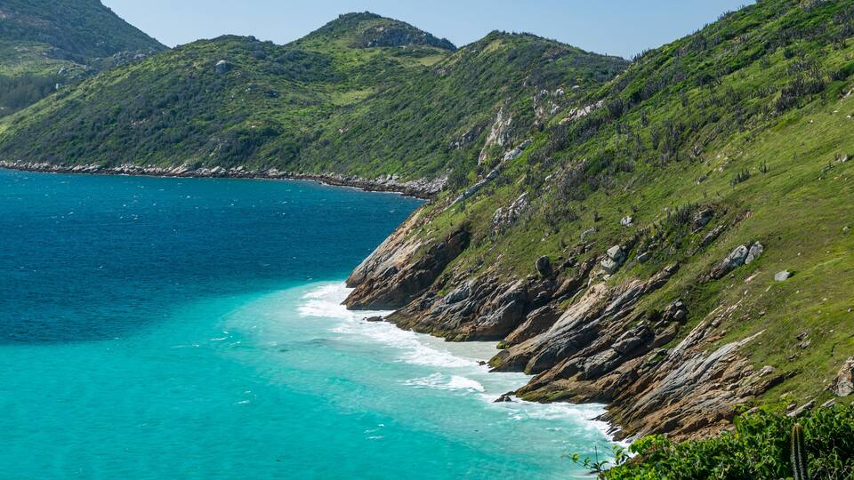A coastal landscape with clear turquoise waters, rocky cliffs, and lush green hills.