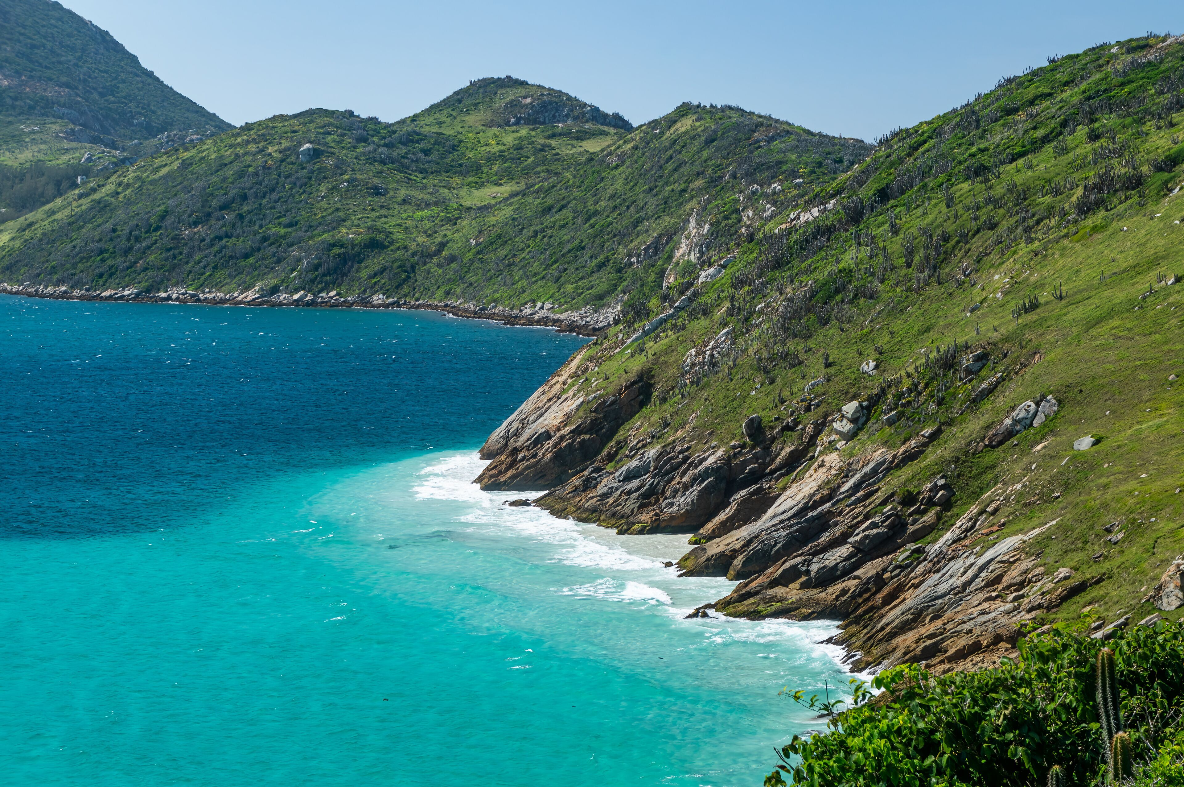 A coastal landscape with clear turquoise waters, rocky cliffs, and lush green hills.