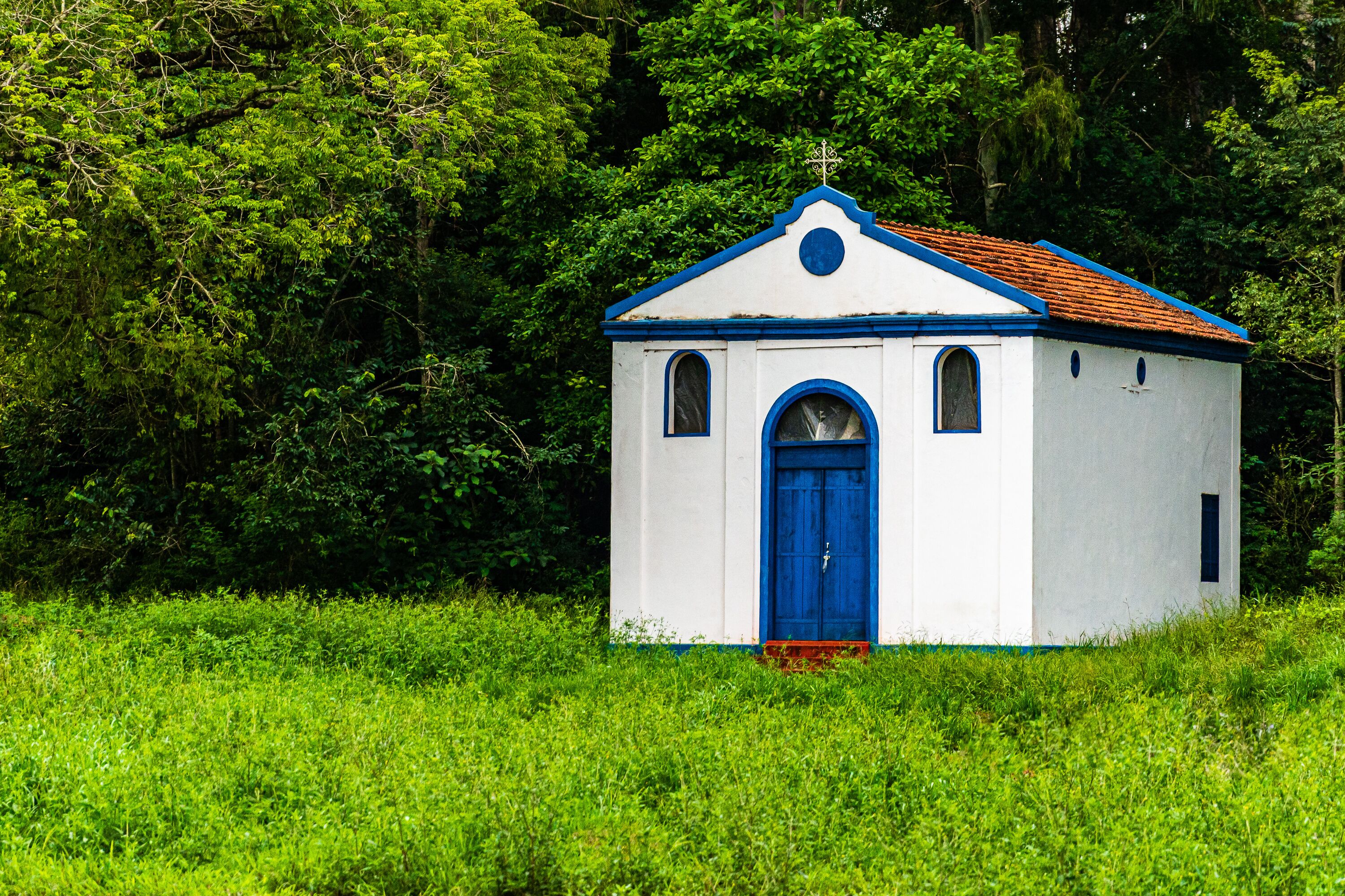 A small white church with a blue door and roof, surrounded by lush green trees.