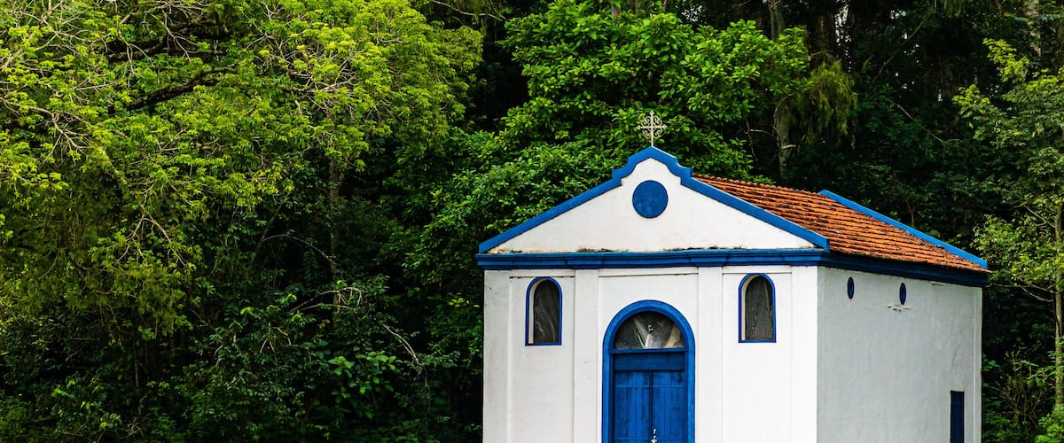 A small white church with a blue door and roof, surrounded by lush green trees.