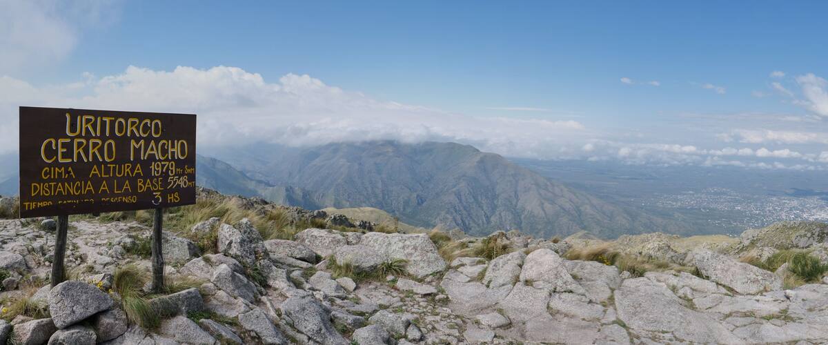 A sign on a mountain peak with a view of a valley below.