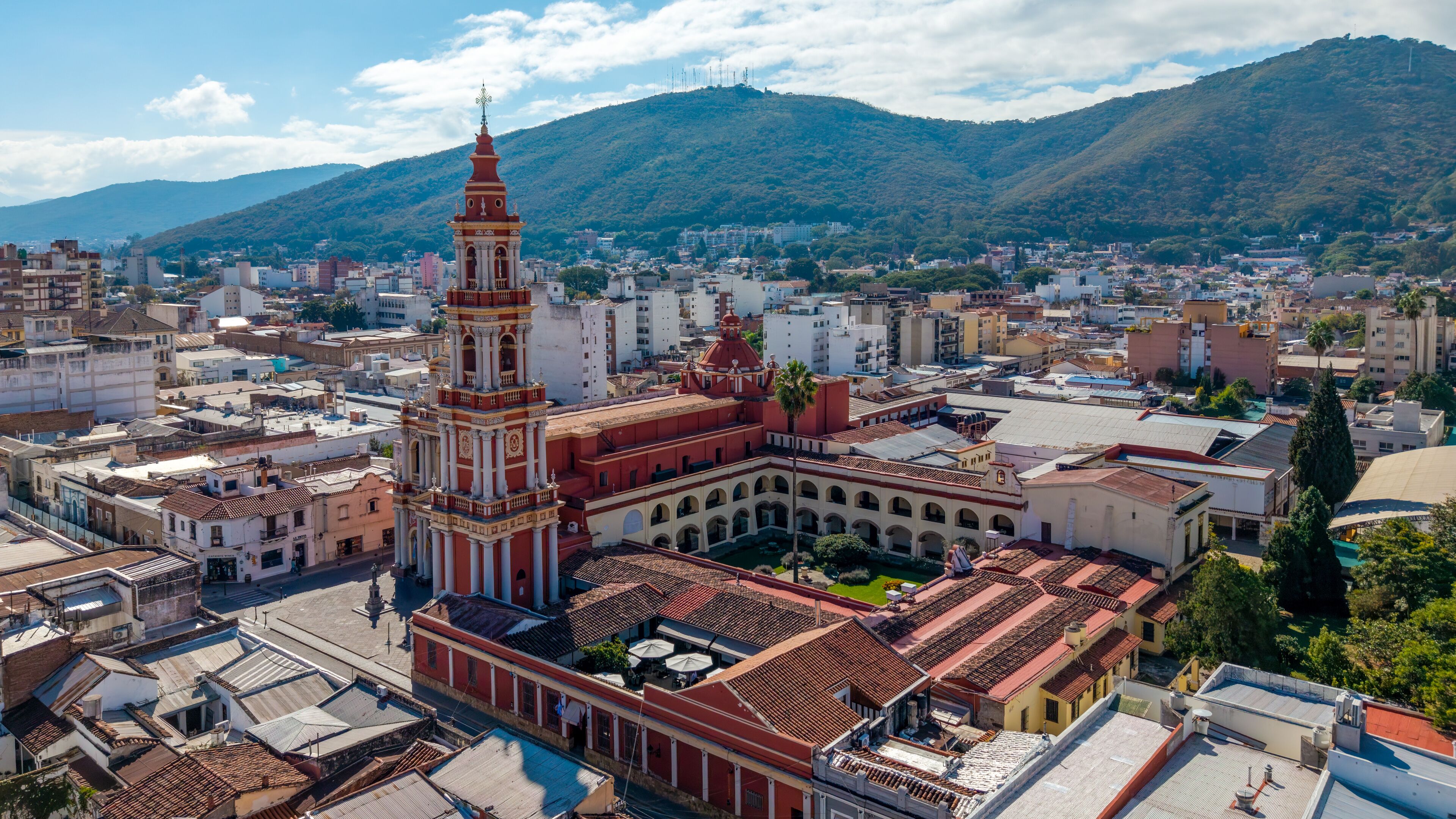 A historic plaza with a prominent clock tower, surrounded by traditional buildings and a mountainous backdrop.