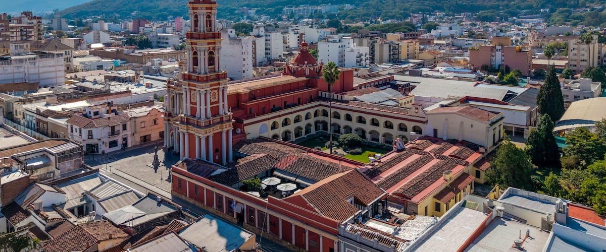 A historic plaza with a prominent clock tower, surrounded by traditional buildings and a mountainous backdrop.