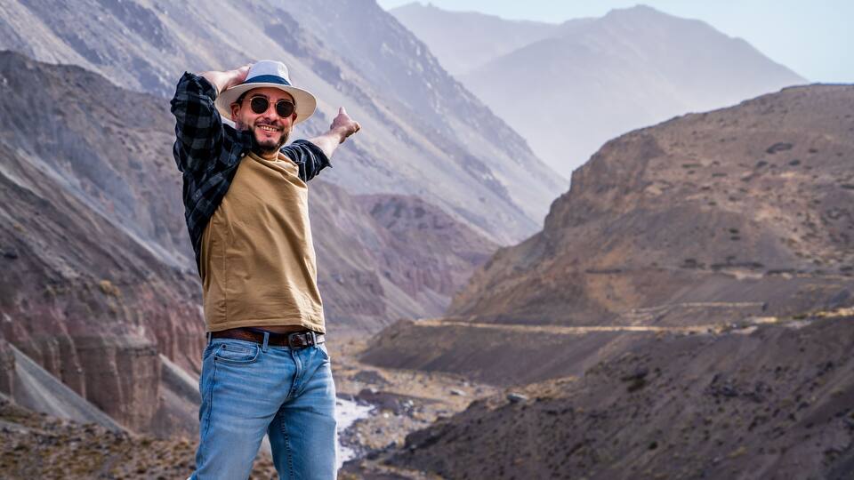 A person in a hat and sunglasses posing in a mountainous landscape with a valley and distant mountains.