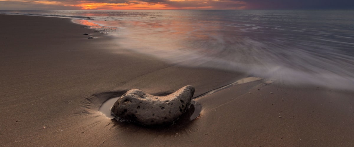 A sandy beach with a large rock in the foreground and a sunset over the ocean.