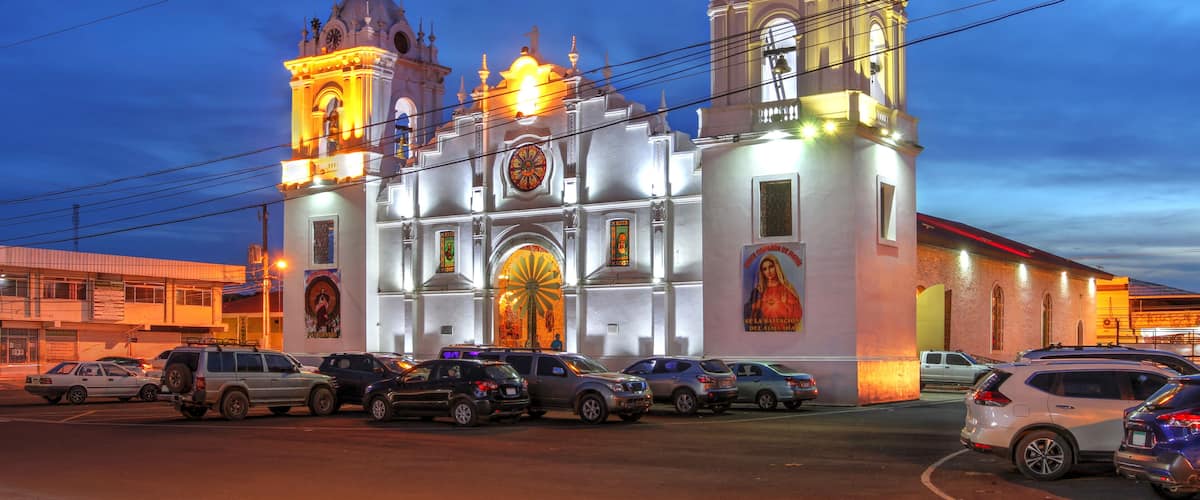 Une église historique, dotée de deux tours et d’un grand dôme central, entourée de voitures garées et bordée par une rue.
