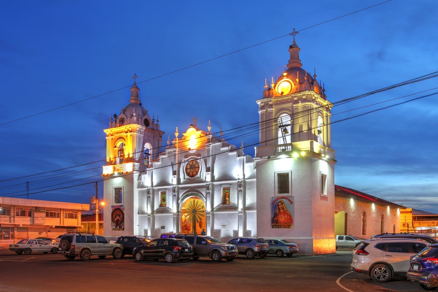 Une église historique, dotée de deux tours et d’un grand dôme central, entourée de voitures garées et bordée par une rue.
