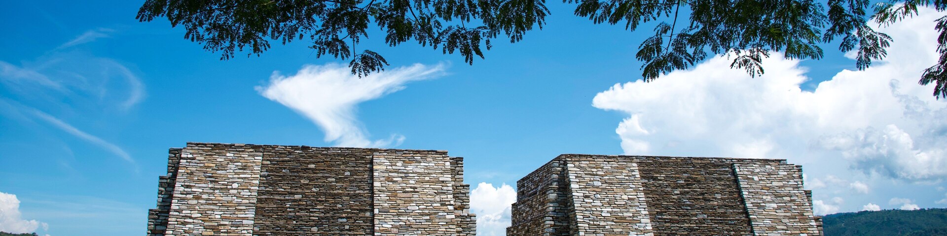 Dos pirámides de piedra antiguas en un campo cubierto de hierba, con un cielo azul despejado y montañas al fondo.