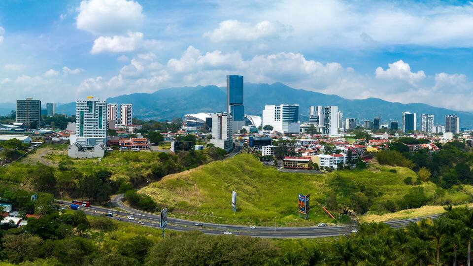 A cityscape with modern buildings, a highway, and green hills under a clear sky.