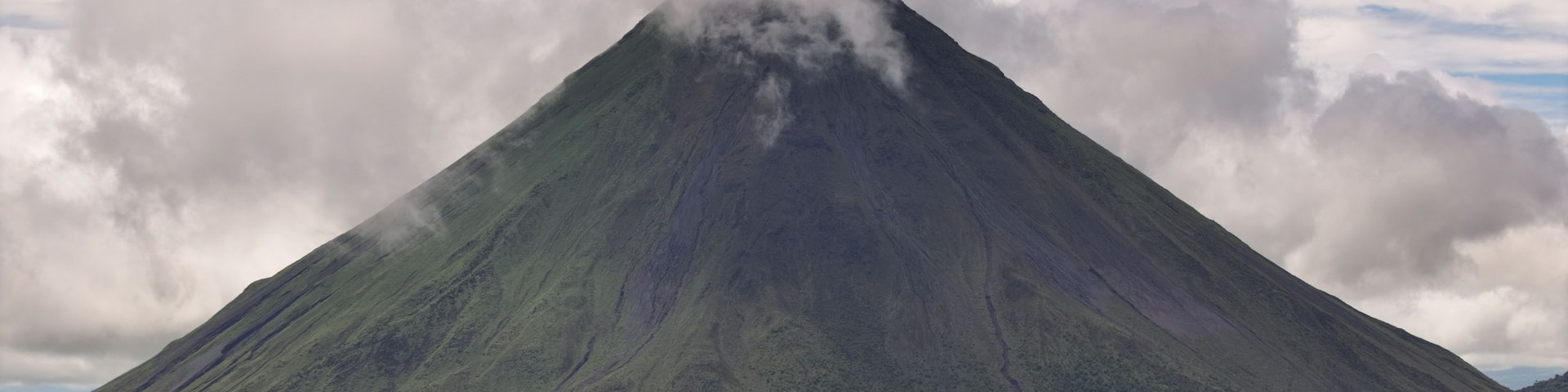 A large, steep-sided volcano with a dense cloud of smoke at its peak, surrounded by a forested landscape.