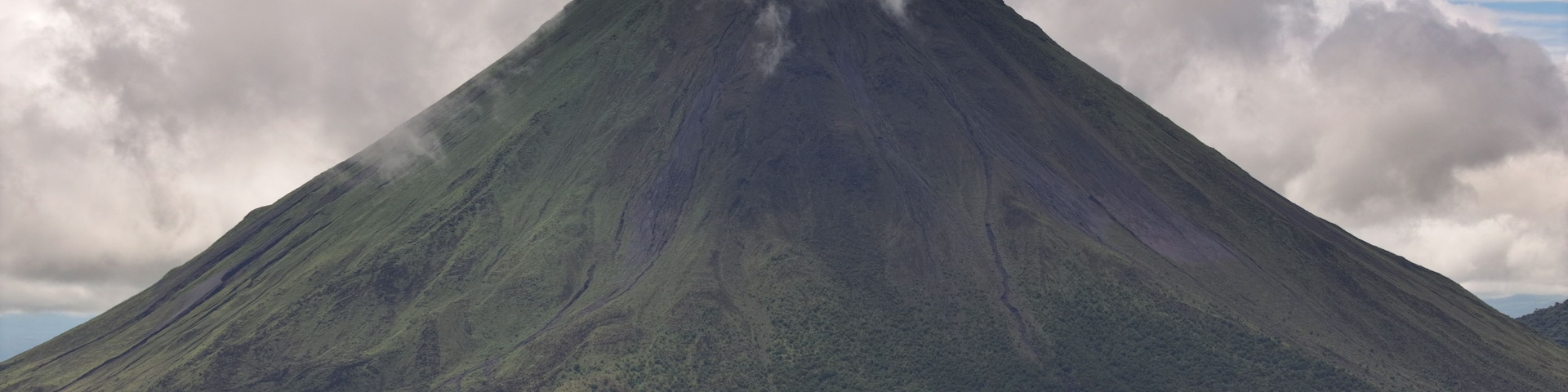 A large, steep-sided volcano with a dense cloud of smoke at its peak, surrounded by a forested landscape.
