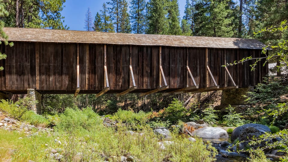 A wooden covered bridge in a forested area with a stream below.