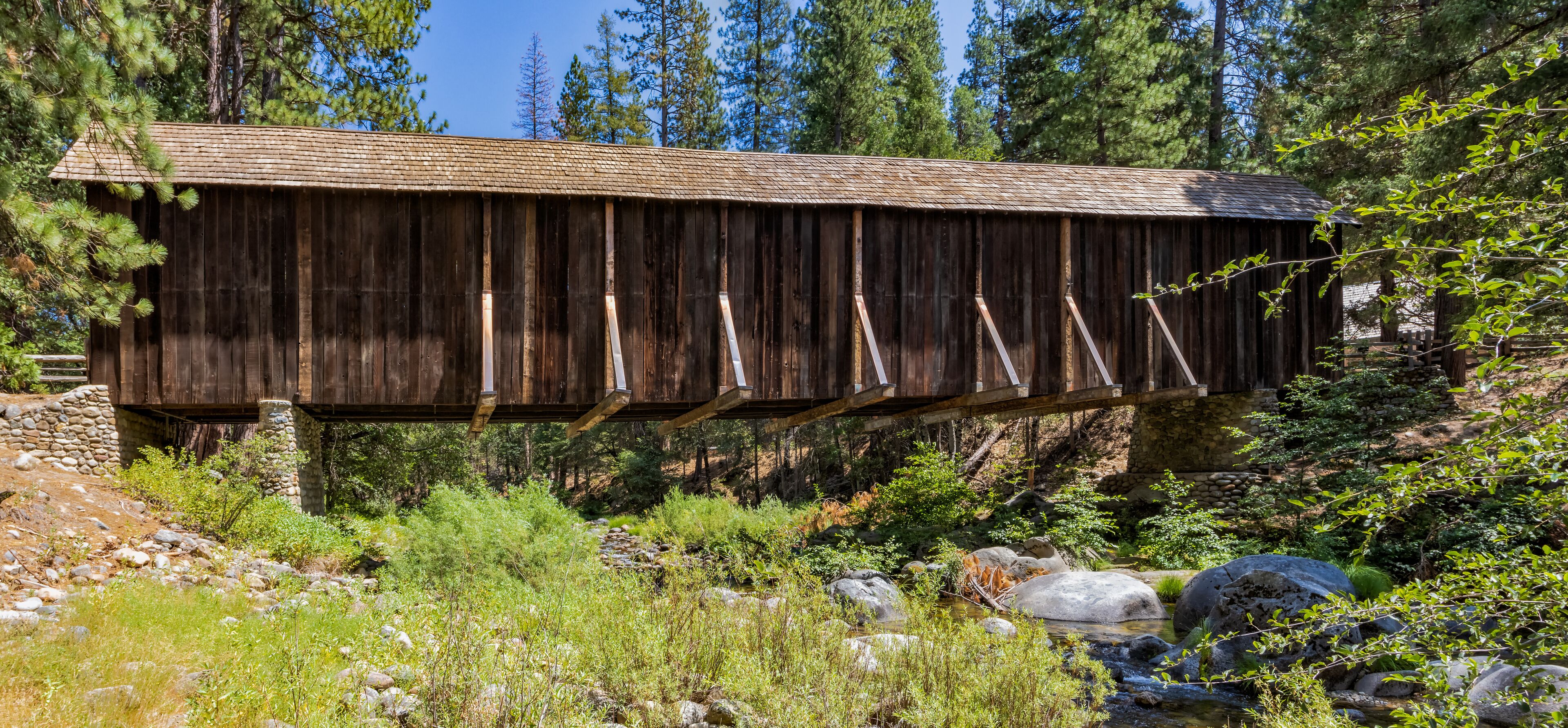 A wooden covered bridge in a forested area with a stream below.