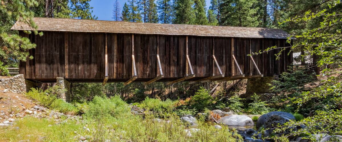 A wooden covered bridge in a forested area with a stream below.
