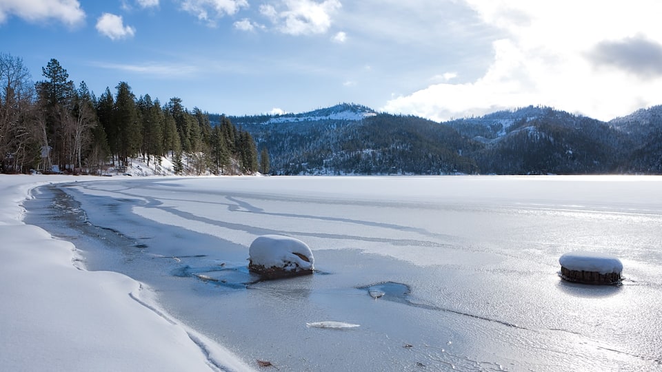 A frozen lake with snow-covered rocks and trees in the background.