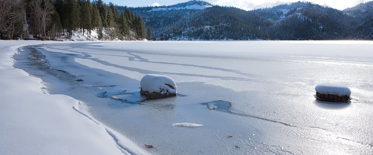 A frozen lake with snow-covered rocks and trees in the background.