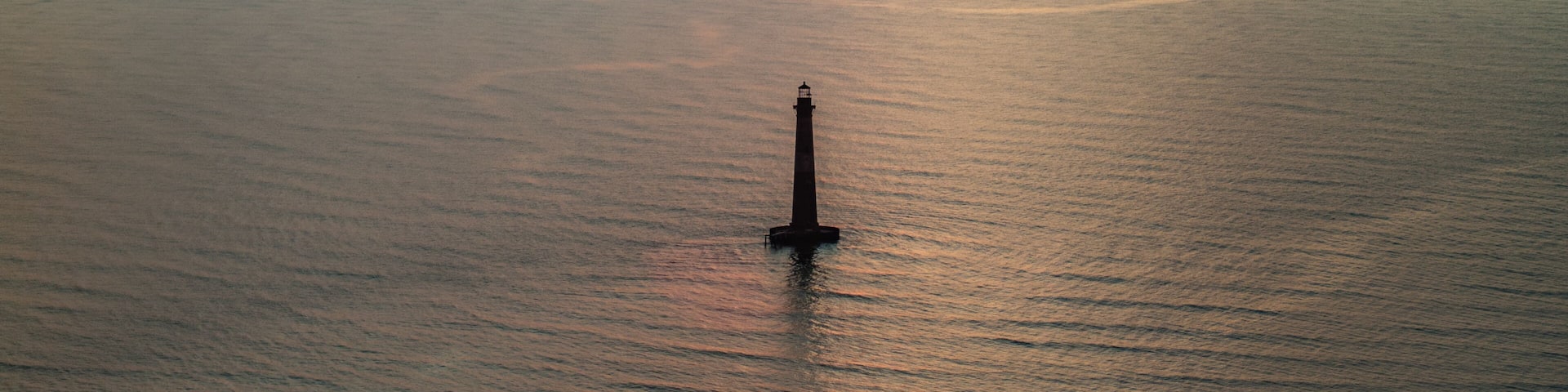 Panormic aerial view of ocean and Morris Island Lighthouse in Folly Beach near Charleston, South Carolina during morning sunrise above horizon over calm waters and golden skies landscape seascape