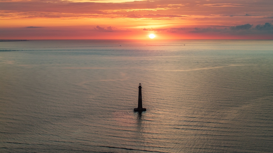 Panormic aerial view of ocean and Morris Island Lighthouse in Folly Beach near Charleston, South Carolina during morning sunrise above horizon over calm waters and golden skies landscape seascape