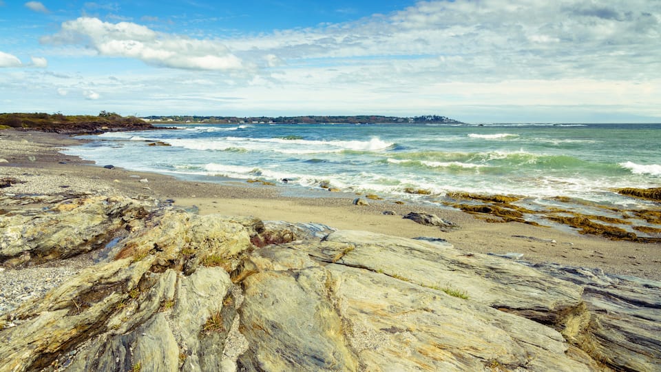 A rocky shoreline with waves crashing onto the beach, a distant shoreline with buildings, and a partly cloudy sky.