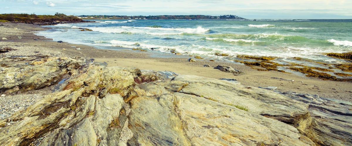 A rocky shoreline with waves crashing onto the beach, a distant shoreline with buildings, and a partly cloudy sky.