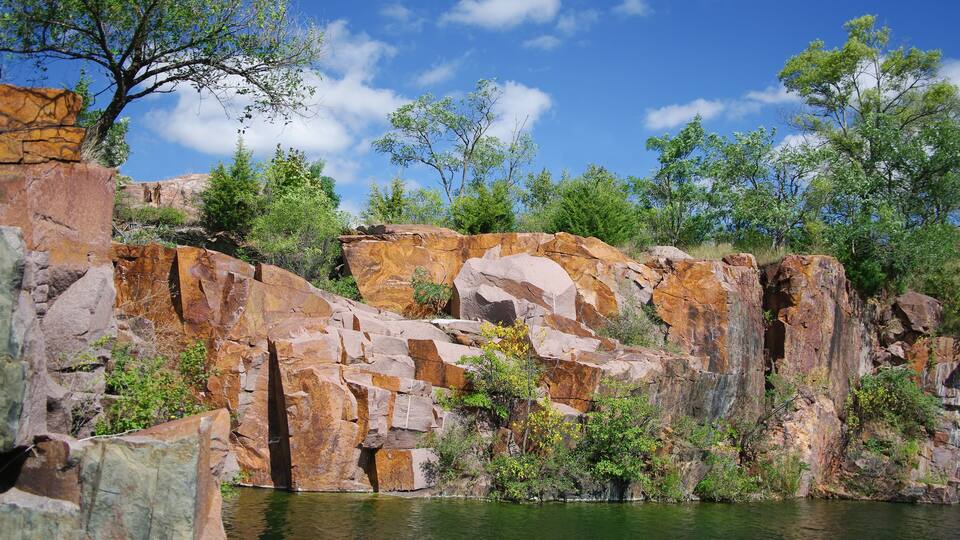 A rocky cliff with a river at its base, surrounded by greenery and trees.